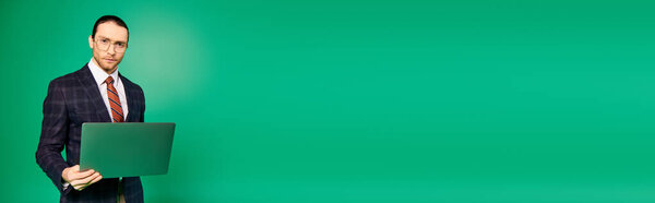 Handsome businessman in a chic suit holding a laptop on a green backdrop.