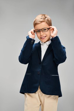 Küçük, şık giyimli, gözlüklü bir çocuk gri bir arkaplana karşı düşünceli bir poz veriyor..