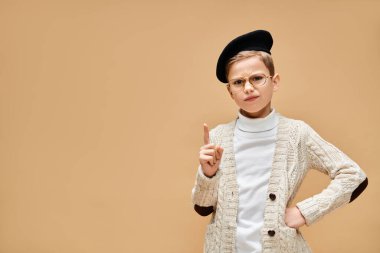 Preadolescent boy in glasses and hat poses as a film director on beige backdrop. clipart