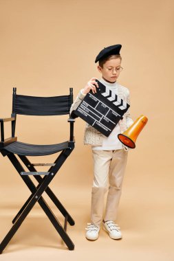 A cute preadolescent boy holding a movie clapper near a chair. clipart