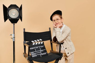 Preadolescent boy dressed as a film director stands next to a camera. clipart