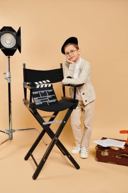 Film yönetmeni gibi giyinmiş ergenlik öncesi bir çocuk yönetmen koltuğunun yanında güvenle durur..