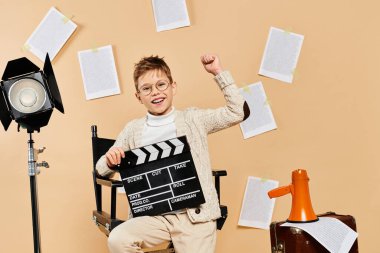 A preadolescent boy dressed as a film director sits with a movie clapper on a beige backdrop. clipart