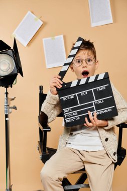 Young boy in film director attire, holding movie clapper in chair. clipart