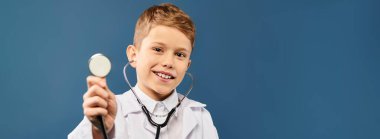 Genç çocuk şakacı bir şekilde doktor gibi davranıyor, elinde steteskop, mavi bir arka planda..