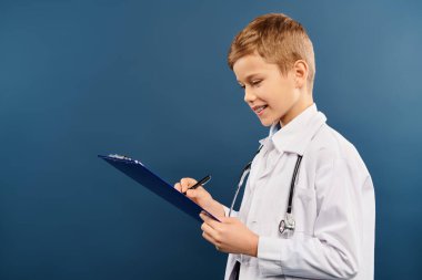 Young boy in white doctors shirt jotting notes on clipboard against blue backdrop. clipart