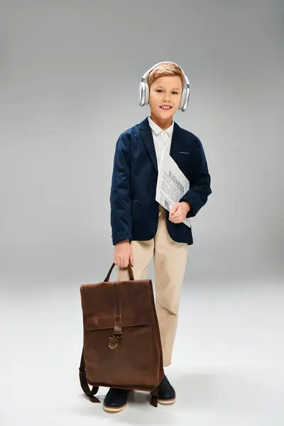 stock image Preadolescent boy in elegant attire, wearing headphones, holds a briefcase on gray backdrop.