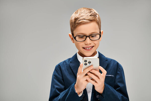 Мальчик в очках, с мобильным телефоном, элегантно одетый на сером фоне.
