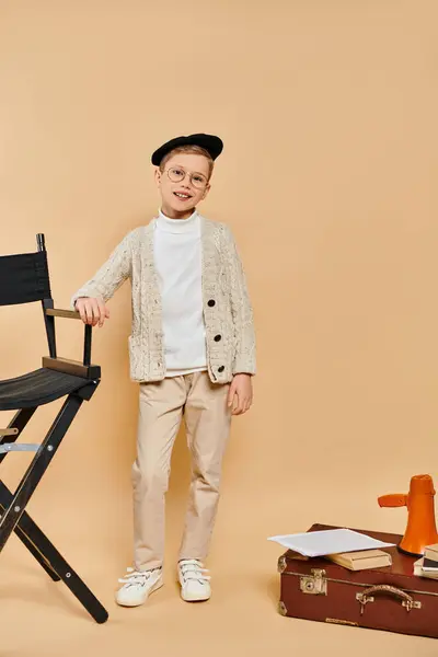 映画監督として服を着たかわいい若い少年は ベージュの背景に椅子の隣に立っています — ストック写真