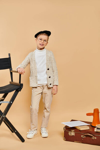 A cute preadolescent boy, dressed as a film director, stands next to a chair on a beige backdrop.