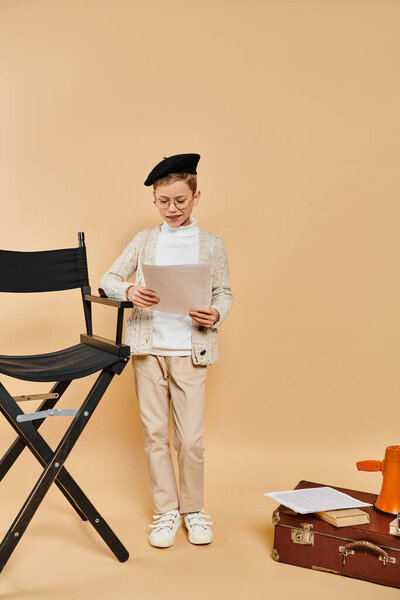 Preadolescent boy in film director costume holds paper next to chair.