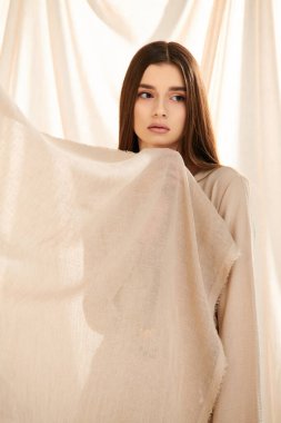 A young woman with long brunette hair strikes a pose in a summer outfit, exuding elegance and beauty in front of a white curtain. clipart