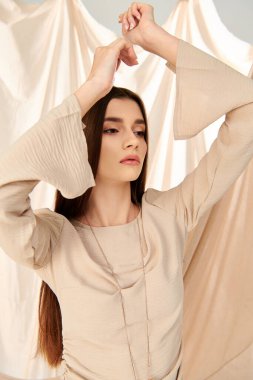 Uzun esmer saçlı genç bir kadın yaz kıyafetiyle poz veriyor. Akan bir perdenin önünde zarif bir şekilde duruyor..