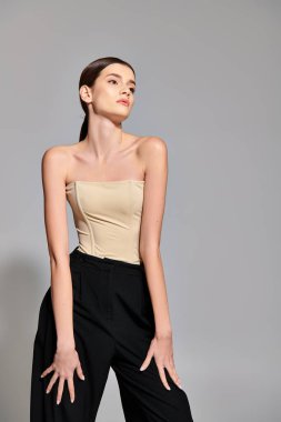 A stylish young woman with brunette hair strikes a pose in a studio setting, showcasing a trendy tan top paired with classic black pants. clipart