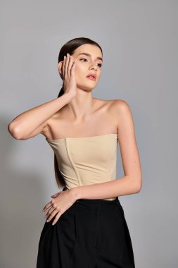 A young woman with brunette hair is elegantly posing in a studio setting, wearing a tan top and black pants. clipart