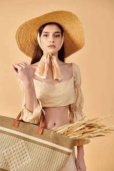 stock image A young brunette woman exudes summer elegance, donning a straw hat and holding a stylish bag in a studio setting.
