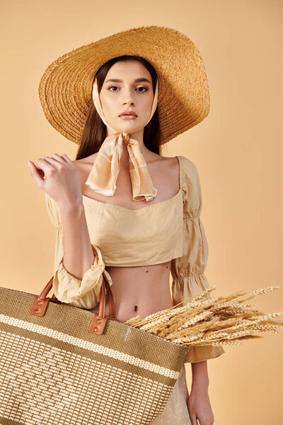 A young brunette woman exudes summer elegance, donning a straw hat and holding a stylish bag in a studio setting.