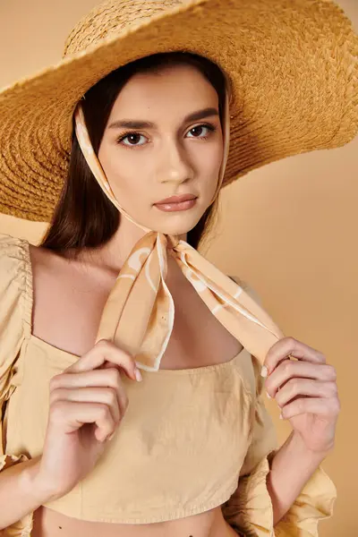 A young woman with long brunette hair poses in a studio setting, exuding a summer vibe in her straw hat and flowing dress.