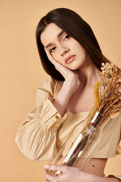 A young woman with long brunette hair holds a vase filled with dried flowers, exuding a serene and peaceful vibe in a summer studio setting.