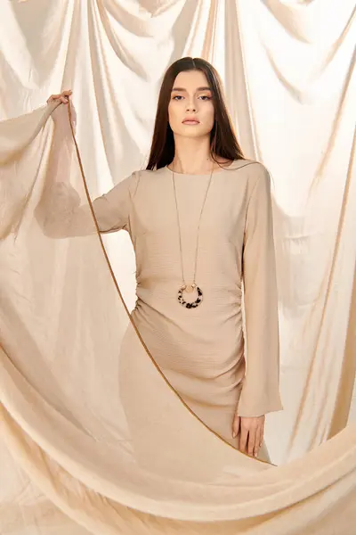 stock image A young woman with long brunette hair poses in a summer outfit in front of a white curtain, exuding serenity and grace.