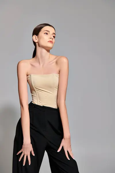 stock image A stylish young woman with brunette hair strikes a pose in a studio setting, showcasing a trendy tan top paired with classic black pants.