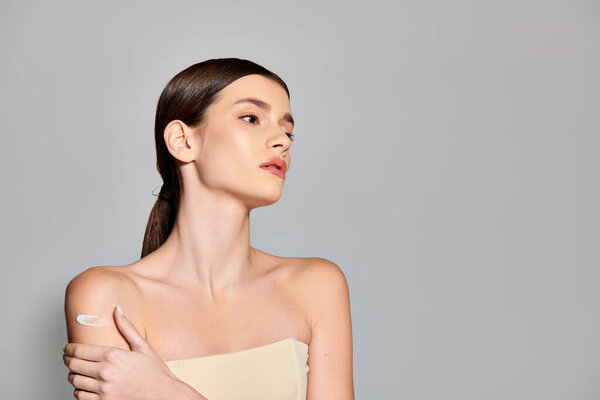 A young woman with brunette hair poses in a strapless dress, gently hugging her own shoulder.