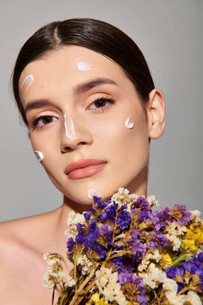 A young woman with brunette hair holds a bouquet of flowers with cream on her face, exuding ethereal beauty.