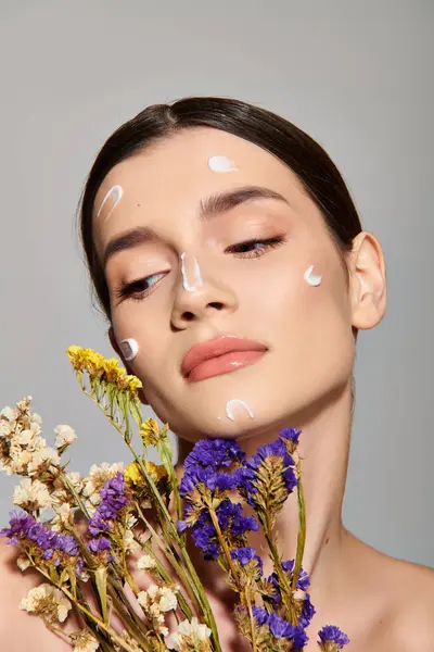 Stock image A young woman with brunette hair holding a bouquet of flowers, her face adorned with white cream, exuding elegance and beauty.