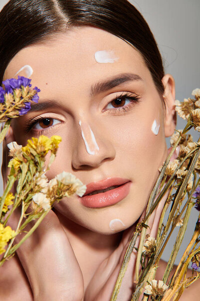 A young woman with brunette hair elegantly holds colorful flowers in front of her face in a studio setting on a grey background.