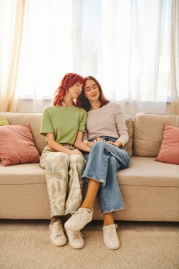 Ortaklık, genç ve memnun lgbt çifti gündelik giysiler içinde el ele tutuşup kanepede oturuyorlar.