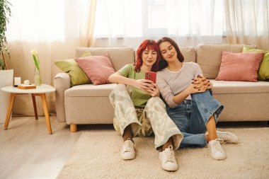 Mutlu lezbiyen çift gülüyor ve akıllı telefonu oturma odasında, modern yaşam tarzında ve sosyal medyada kullanıyor.