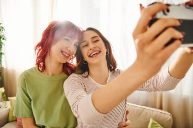 Genç ve gülümseyen lezbiyen çift geçmişe dönük kamerayla selfie çekiyorlar, evlerinde mutlu anları yakalıyorlar.