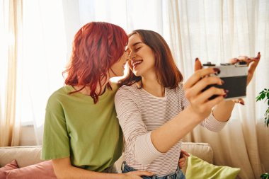 young lesbian couple smiling and taking selfie on retro camera, capturing blissful moment at home clipart