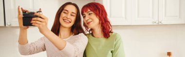 banner of young lesbian couple taking selfie on retro camera in kitchen, capturing happy moment clipart