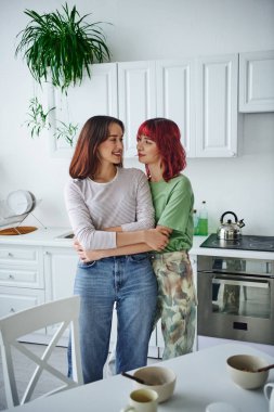 Modern mutfakta kız arkadaşını kucaklayan kızıl saçlı, mutlu ve piercingli lezbiyen kadın.