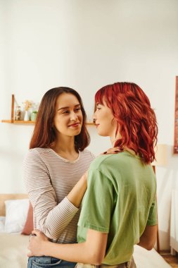 tender young lesbian woman hugging her happy girlfriend with red hair in modern apartment clipart