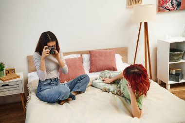 home photo session of lesbian woman taking photo on retro camera of her girlfriend on bed clipart