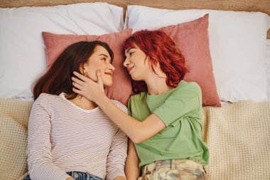 top view of smiling lesbian couple looking at each other and lying together on bed, touching face clipart