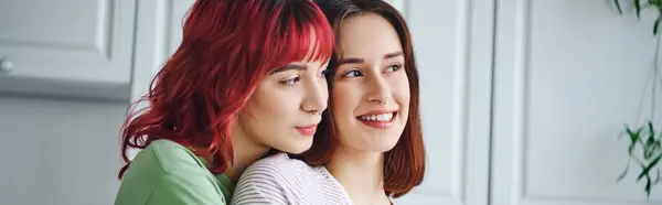 stock image portrait of joyful and pierced lesbian woman with red hair embracing her girlfriend at home, banner