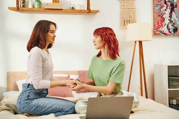 stock image happy young lesbian women holding hands and sitting together on bed next to laptop, lgbt couple