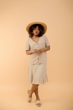 Güzel, kıvırcık saçlı, çiçekli bir yaz elbisesi ve şık, geniş kenarlı bir şapkayla poz veren Afro-Amerikan bir kadın..