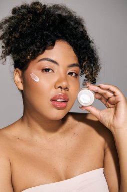 Kıvırcık saçlı güzel bir Afrikalı Amerikalı kadın stüdyo ortamında yüzüne krem sürüyor..