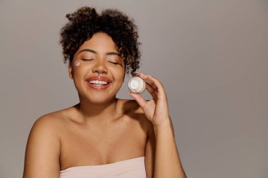 A young African American woman with curly hair smiles as she holds a jar of cream, exuding beauty and confidence. clipart