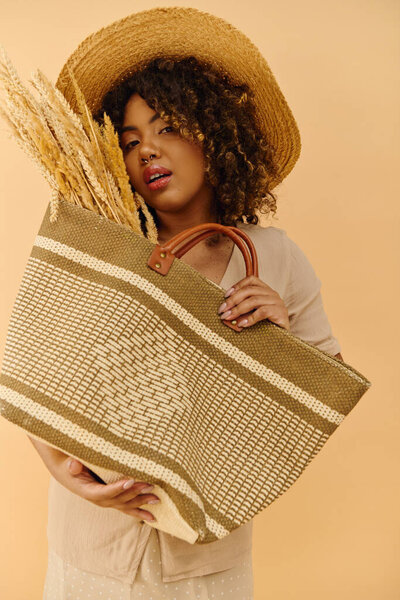 A beautiful young African American woman with curly hair holds a bag with a straw hat on top in a studio setting.