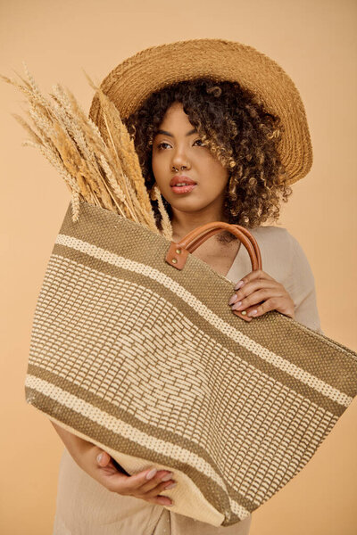 Beautiful African American woman with curly hair in a summer dress holding a bag with a straw hat on top in a studio setting.
