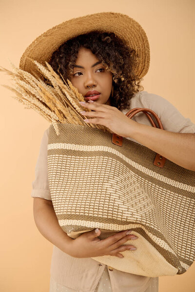A fashionable African American woman with curly hair poses in a studio, holding a bag while sporting a straw hat.