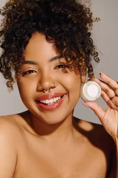 stock image Curly-haired African American woman holding a jar of cream, showcasing skin care routine in a studio setting.