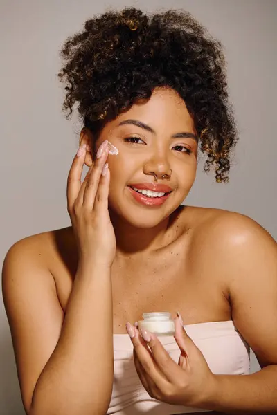 stock image Beautiful African American woman with curly hair holding a jar of cream in front of her face in a studio setting.