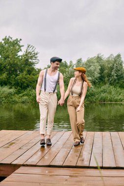 Bir adam ve bir kadın, gelişigüzel giyinmiş, yemyeşil bir parkta kırsal ahşap bir köprüde el ele dolaşıyorlar..