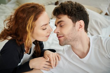 Bir adam ve bir kadın yan yana yatıyorlar, gün ışığının yumuşak parıltısı altında samimi bir anı paylaşıyorlar..